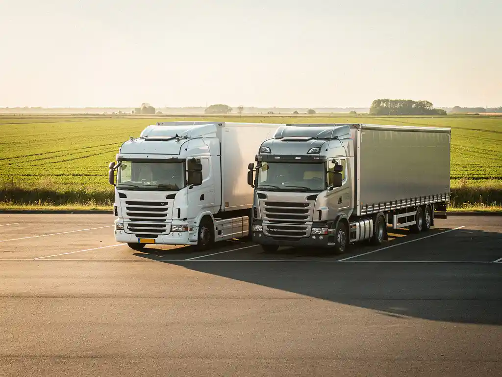 Witte eigen vrachtwagen en externe logistieke carrier zij aan zij geparkeerd op Nederlands snelwegparkeerplaats bij gouden uurlicht.